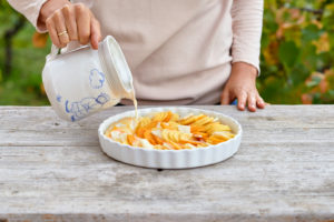 Vorbereitung eines Gratin von Kartoffeln, Kürbis und Topinambur, Übergießen der aufgeschnittenen Zutaten in einer gebutterten Backform mit Milch und Sahne aus einer Kanne Vorbereitung eines Gratin von Kartoffeln, Kürbis und Topinambur, Übergießen der aufgeschnittenen Zutaten in einer gebutterten Backform mit Milch und Sahne aus einer Kanne